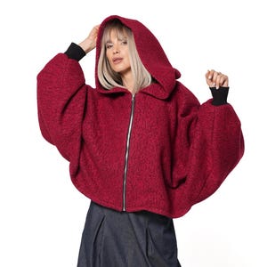Puede incluir: Una chaqueta con capucha y cremallera de color rojo oscuro, texturizada, con mangas anchas y puños acanalados negros, combinada con pantalones azul oscuro plisados de corte holgado y botas de plataforma negras. La chaqueta tiene cremallera.