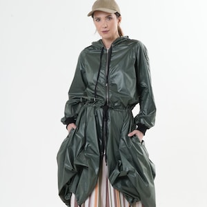 Puede incluir: Una chaqueta verde con capucha, cintura con cordón y cierre de cremallera. La chaqueta está hecha de un material brillante y resistente al agua. La modelo lleva la chaqueta con un pantalón a rayas.