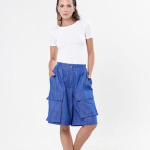 May include: A woman wearing a white short-sleeve t-shirt and blue shorts with large pockets. The shorts are knee-length. She is wearing brown sandals with a platform sole. The background is white.