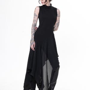 May include: A black sleeveless dress featuring a high neckline and an asymmetrical pleated skirt. The dress is worn with black open-toe boots. The model is standing against a white backdrop.