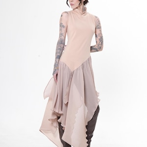 Op de afbeelding: Een beige mouwloze jurk met een hoge halslijn en een geplooide, asymmetrische rok. De jurk wordt gecombineerd met zwarte open-teen hakken en bruine leren laarzen. De algehele stijl is elegant en modern, geschikt voor een speciale gelegenheid.