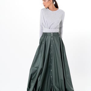 Op de afbeelding: Een lange, donkergroene rok met een knoopsluiting aan de voorkant en een volumineus, geplooid ontwerp. De rok wordt gecombineerd met een lichtgrijze top met lange mouwen. De outfit staat tegen een witte achtergrond.