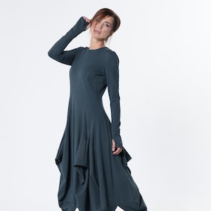 May include: A dark teal long-sleeved dress with an unusual asymmetrical hem. The dress has a round neckline and fitted sleeves. The model is wearing light-coloured lace-up boots. The dress is made of a soft, flowing fabric.