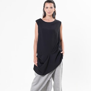 Könnte beinhalten: Eine Frau trägt ein schwarzes ärmelloses Oberteil mit einem drapierten Design und graue Leinenhose. Das Outfit umfasst schwarze Sandalen. Der Hintergrund ist schlicht weiß.