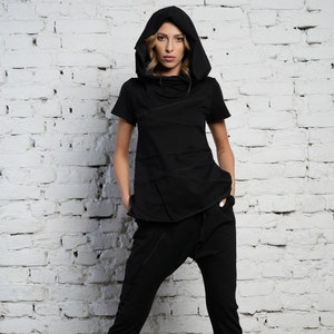 May include: A black hooded top with a layered design and a matching pair of black pants. The model is wearing the outfit with white sandals.