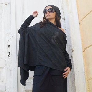 Könnte beinhalten: Eine Frau trägt einen dunkelgrauen, übergroßen Pullover mit einer schwarzen Mütze und einer Sonnenbrille. Sie trägt eine schwarze Hose mit lockerer Passform und schwarze Sneaker mit weißem Text, der "Always Be Kind" lautet.