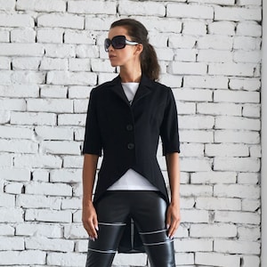 May include: A black blazer with a long tail, short sleeves, and three buttons. The blazer is worn over a white t-shirt and black leather pants with white stitching. The person is wearing black sunglasses and black heeled boots.