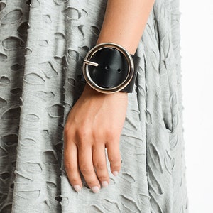 Puede incluir: Un brazalete de cuero negro con una hebilla circular grande de plata.