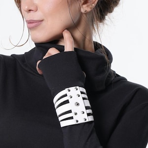 Może przedstawiać: Czarny golf z długim rękawem z białym mankietem z czarnymi paskami i srebrnymi ćwiekami. Top jest połączony z szarą spódnicą. Modelka ma na sobie top i spódnicę na białym tle.