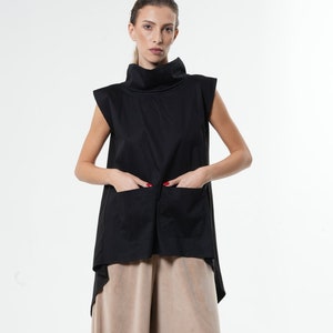 Op de afbeelding: Een zwart, mouwloos top met een hoge, ronde hals en een asymmetrische zoom met twee zakken. Het top wordt gecombineerd met een wijde, lichtbruine broek.