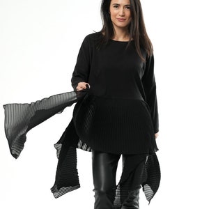 Puede incluir: Una blusa negra de manga larga con falda plisada y una estola negra transparente. La blusa tiene un escote redondo y la estola está drapeada sobre los hombros y atada a la cintura.