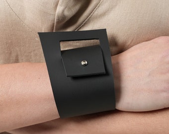 Puede incluir: Un brazalete de cuero negro con una hebilla cuadrada y una solapa pequeña. La hebilla tiene un remache plateado.