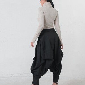 May include: A woman wearing a beige turtleneck sweater and black, loose-fitting pants with a gathered design. She is wearing black heeled sandals.