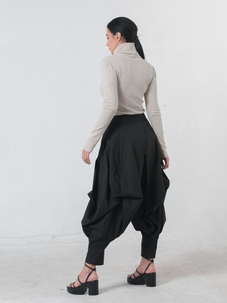 May include: A woman wearing a beige turtleneck sweater and black, loose-fitting pants with a gathered design. She is wearing black heeled sandals.