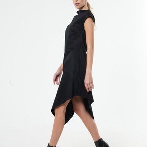 K&ouml;nnte beinhalten: Ein schwarzes asymmetrisches Kleid mit Stehkragen und kurzen &Auml;rmeln. Das Kleid ist am Oberk&ouml;rper figurbetont und flie&szlig;t in einen l&auml;ngeren, asymmetrischen Saum aus. Das Model tr&auml;gt schwarze Stiefeletten mit Absatz.