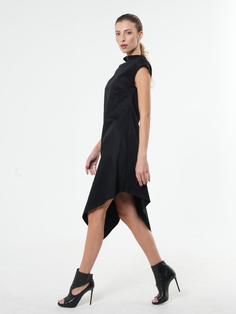 K&ouml;nnte beinhalten: Ein schwarzes asymmetrisches Kleid mit Stehkragen und kurzen &Auml;rmeln. Das Kleid ist am Oberk&ouml;rper figurbetont und flie&szlig;t in einen l&auml;ngeren, asymmetrischen Saum aus. Das Model tr&auml;gt schwarze Stiefeletten mit Absatz.