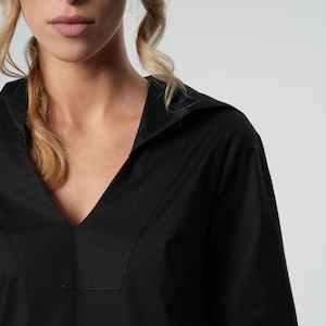 May include: Close-up of a black, long-sleeved top with a V-neck and a collar. The top is made of a smooth fabric and has a pocket detail. The background is plain white.