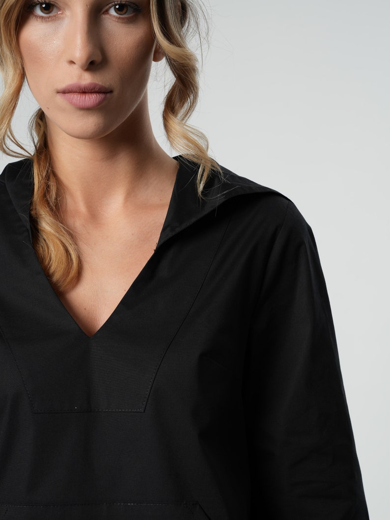 May include: Close-up of a black, long-sleeved top with a V-neck and a collar. The top is made of a smooth fabric and has a pocket detail. The background is plain white.