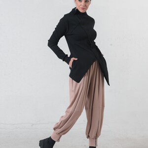 May include: A black mock neck shirt with a long sleeve and a diagonal zipper detail. The shirt is paired with loose-fitting, light brown pants with a zipper detail at the ankles. The model is wearing black ankle boots.