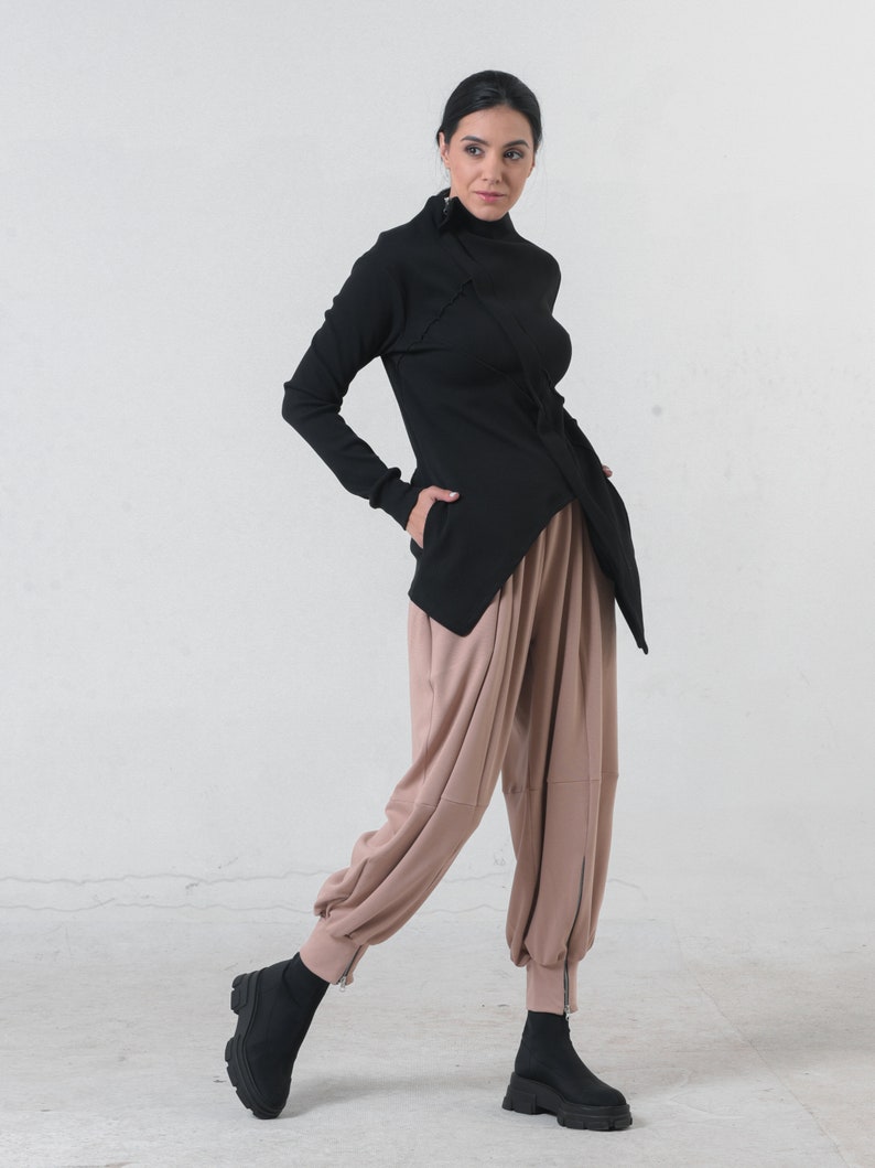 May include: A black mock neck shirt with a long sleeve and a diagonal zipper detail. The shirt is paired with loose-fitting, light brown pants with a zipper detail at the ankles. The model is wearing black ankle boots.