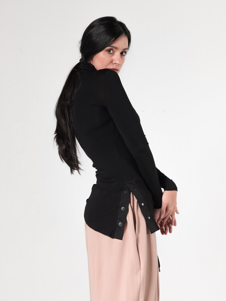 May include: A black long-sleeved shirt with a high neckline and a side panel of buttons. The shirt is tucked into a pair of light brown pants.