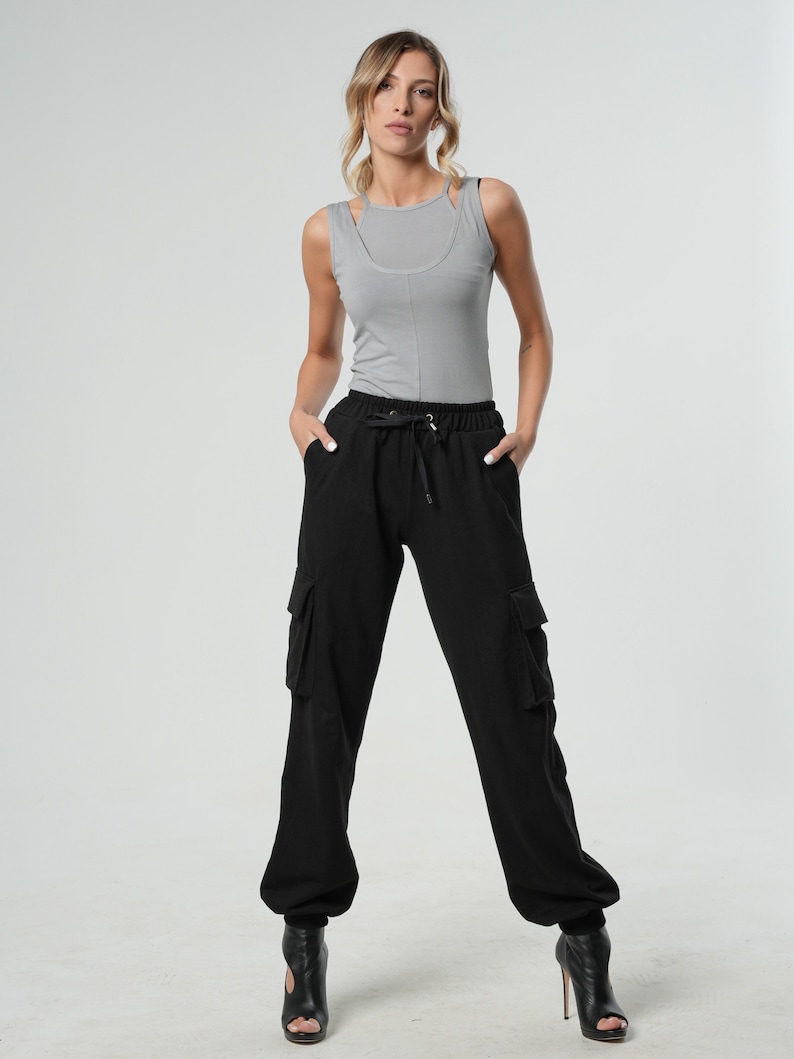 May include: A woman is wearing a gray tank top and black cargo pants with a drawstring waist. She is standing with her hands in her pockets and is wearing black heeled shoes.