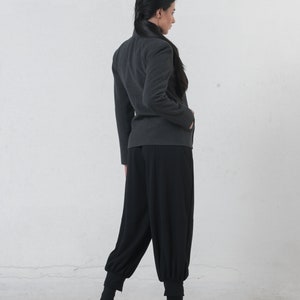 May include: A dark gray jacket with a high collar and long sleeves. The jacket is paired with black, loose-fitting pants that gather at the ankles. The outfit is completed with black ankle boots. The background is a plain white wall.