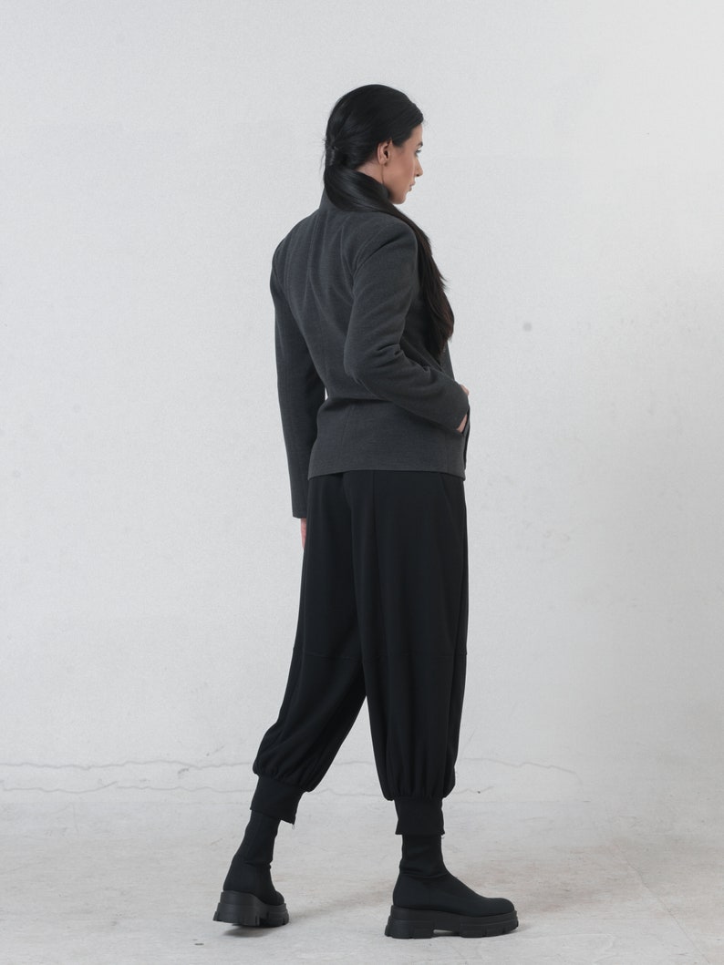 May include: A dark gray jacket with a high collar and long sleeves. The jacket is paired with black, loose-fitting pants that gather at the ankles. The outfit is completed with black ankle boots. The background is a plain white wall.