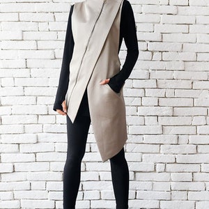 May include: A long, asymmetrical, beige vest with a zipper closure. The vest has a high collar and a single pocket on the left side. The model is wearing black leggings and black heeled boots.