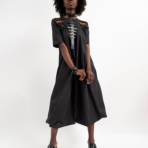 May include: A black midi dress with a cut-out design at the shoulders and a black leather harness with silver accents. The dress has a loose fit and falls to the knees. The model is wearing black platform sandals.