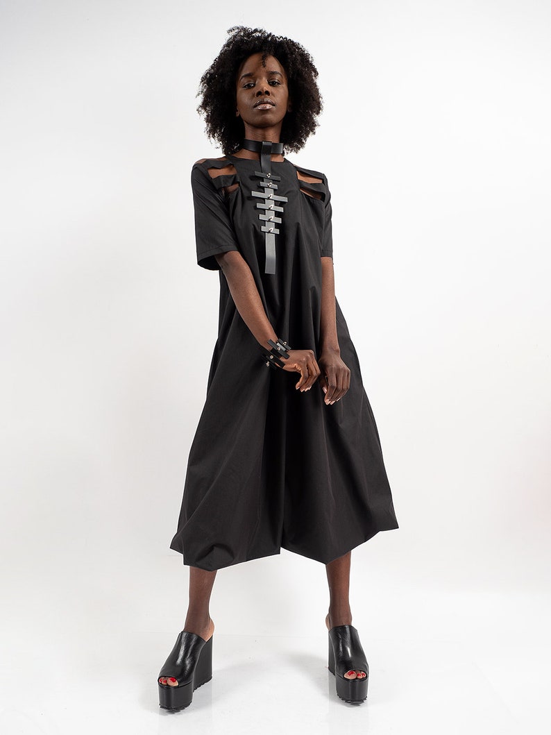 May include: A black midi dress with a cut-out design at the shoulders and a black leather harness with silver accents. The dress has a loose fit and falls to the knees. The model is wearing black platform sandals.