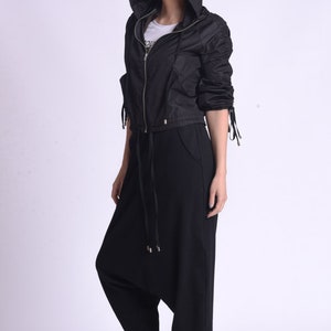 May include: A black hooded jacket with a zipper closure and drawstring waist. The jacket has long sleeves with ties at the cuffs. The model is wearing black pants and black sneakers.