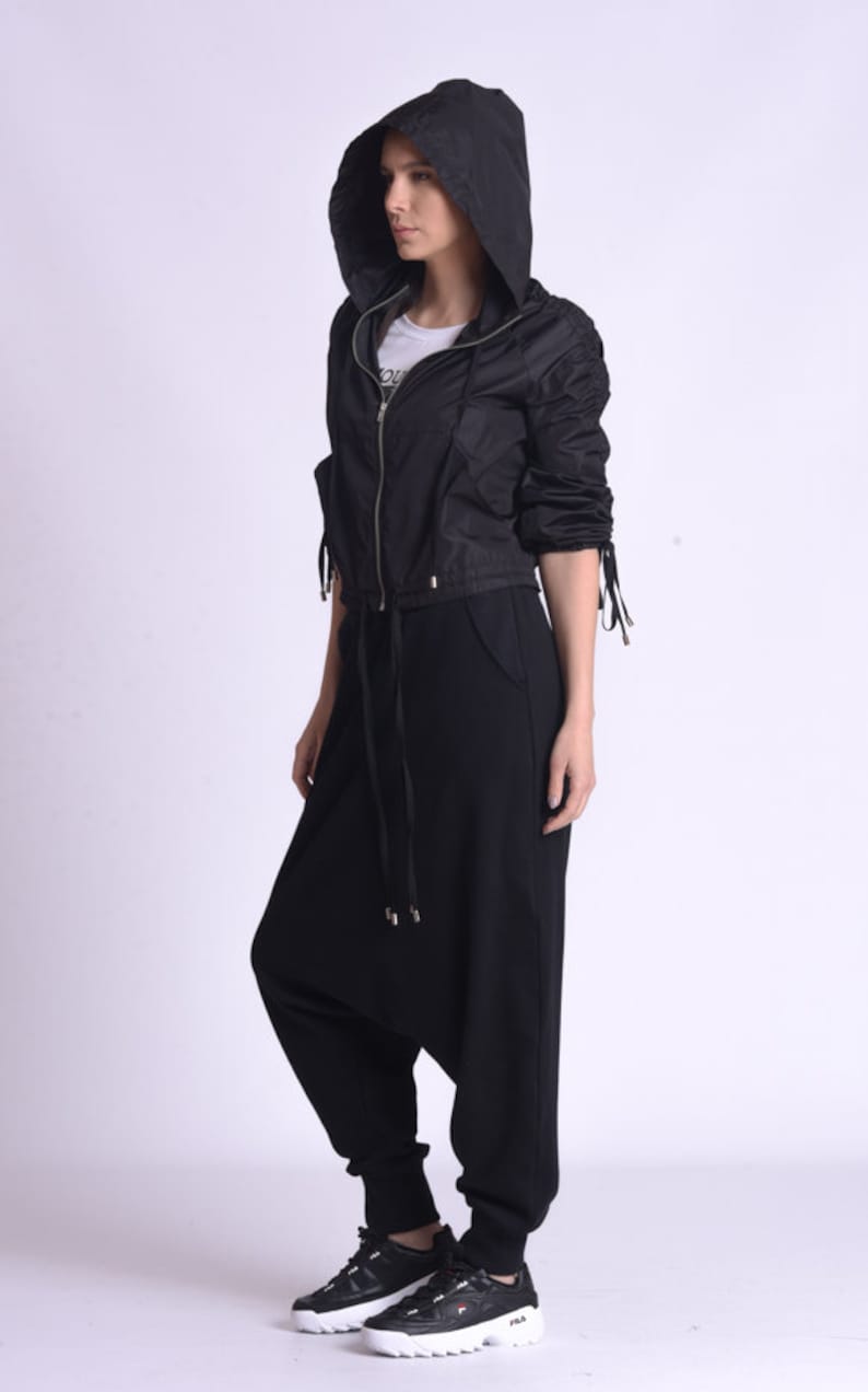 May include: A black hooded jacket with a zipper closure and drawstring waist. The jacket has long sleeves with ties at the cuffs. The model is wearing black pants and black sneakers.
