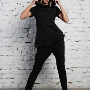 May include: A black hooded top with a short sleeve and a black pair of pants. The model is wearing the outfit and standing in front of a white brick wall.
