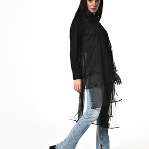 May include: A black long-sleeved shirt with a layered tulle skirt. The shirt has a mock neck and a button-up front. The model is wearing blue jeans and black heeled sandals.