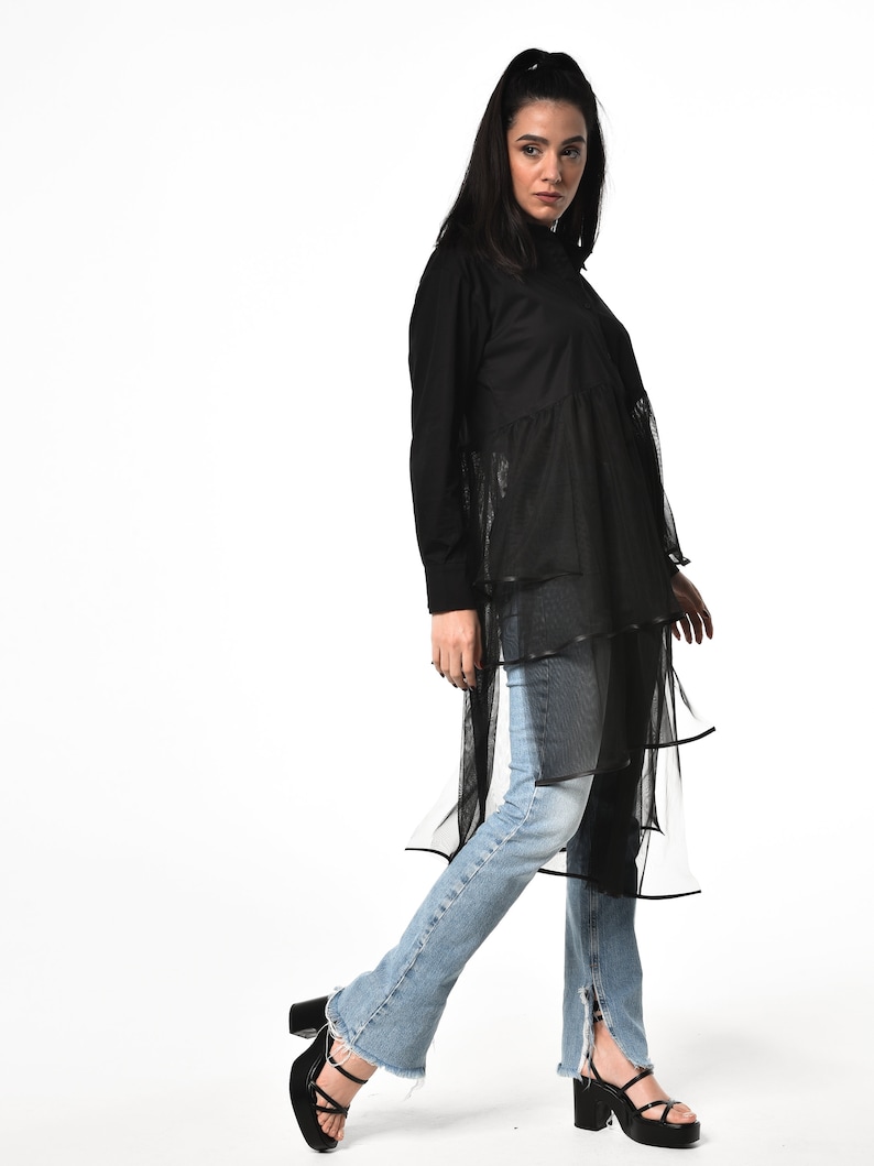 May include: A black long-sleeved shirt with a layered tulle skirt. The shirt has a mock neck and a button-up front. The model is wearing blue jeans and black heeled sandals.