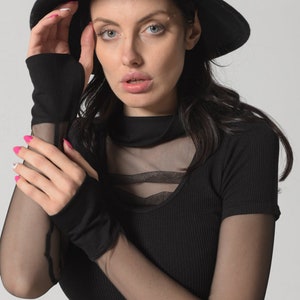 May include: A woman wearing a black fedora hat, a black mesh top with a black ribbed undershirt, and black fingerless gloves.