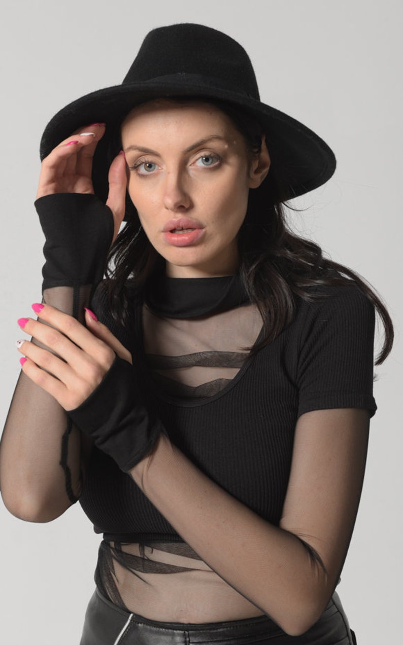 May include: A woman wearing a black fedora hat, a black mesh top with a black ribbed undershirt, and black fingerless gloves.