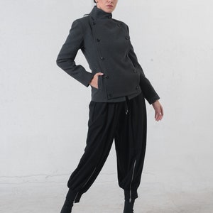 May include: A charcoal gray jacket with a high collar and multiple dark buttons. The jacket is paired with black, loose-fitting pants with zippers at the ankles and black boots. The outfit is a modern, stylish ensemble.