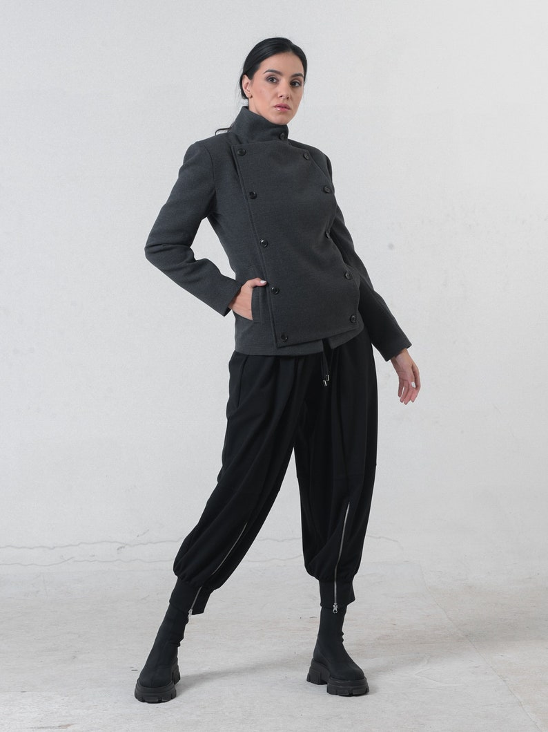 May include: A charcoal gray jacket with a high collar and multiple dark buttons. The jacket is paired with black, loose-fitting pants with zippers at the ankles and black boots. The outfit is a modern, stylish ensemble.