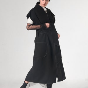 May include: A black long wrap-style vest with a contrasting stitch detail. The vest has a wide collar and a belt that ties at the waist. The model is wearing the vest over a white shirt and black sheer sleeves.