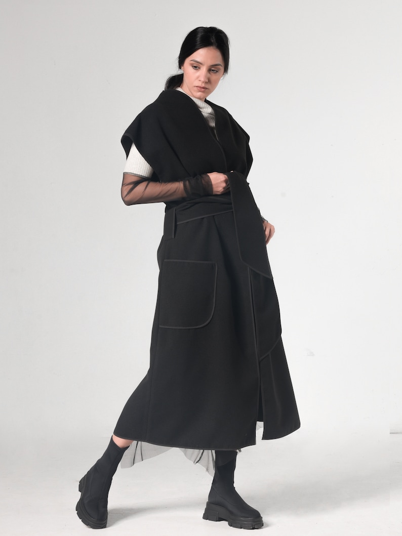 May include: A black long wrap-style vest with a contrasting stitch detail. The vest has a wide collar and a belt that ties at the waist. The model is wearing the vest over a white shirt and black sheer sleeves.