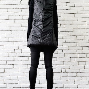 May include: A black puffer vest with a large, dramatic hood. The vest is worn over a black long-sleeved shirt and black pants. The person is standing in front of a white brick wall.