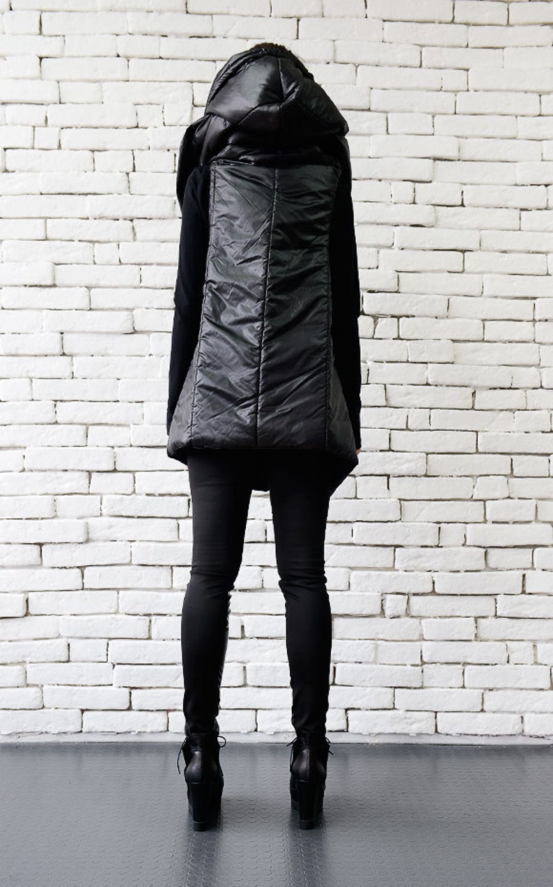 May include: A black puffer vest with a large, dramatic hood. The vest is worn over a black long-sleeved shirt and black pants. The person is standing in front of a white brick wall.