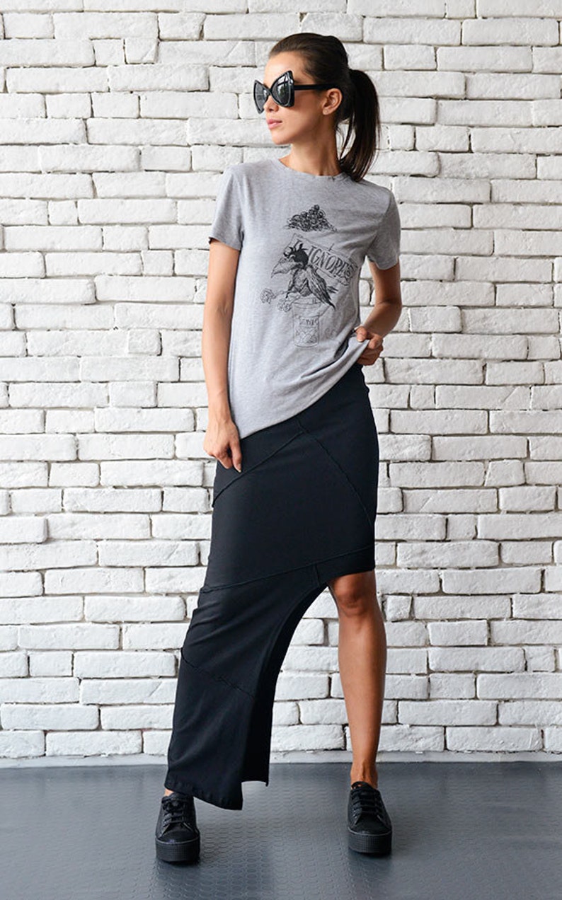 May include: A woman wearing a grey t-shirt with a graphic print of a bird and the word "IGNORES" on it. She is also wearing a black asymmetrical skirt and black platform sneakers.
