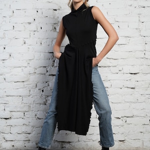 Op de afbeelding: Een zwart shirt met een hoge hals en een lange, asymmetrische zoom. Het shirt wordt gedragen met een blauwe jeans en zwarte hakken.