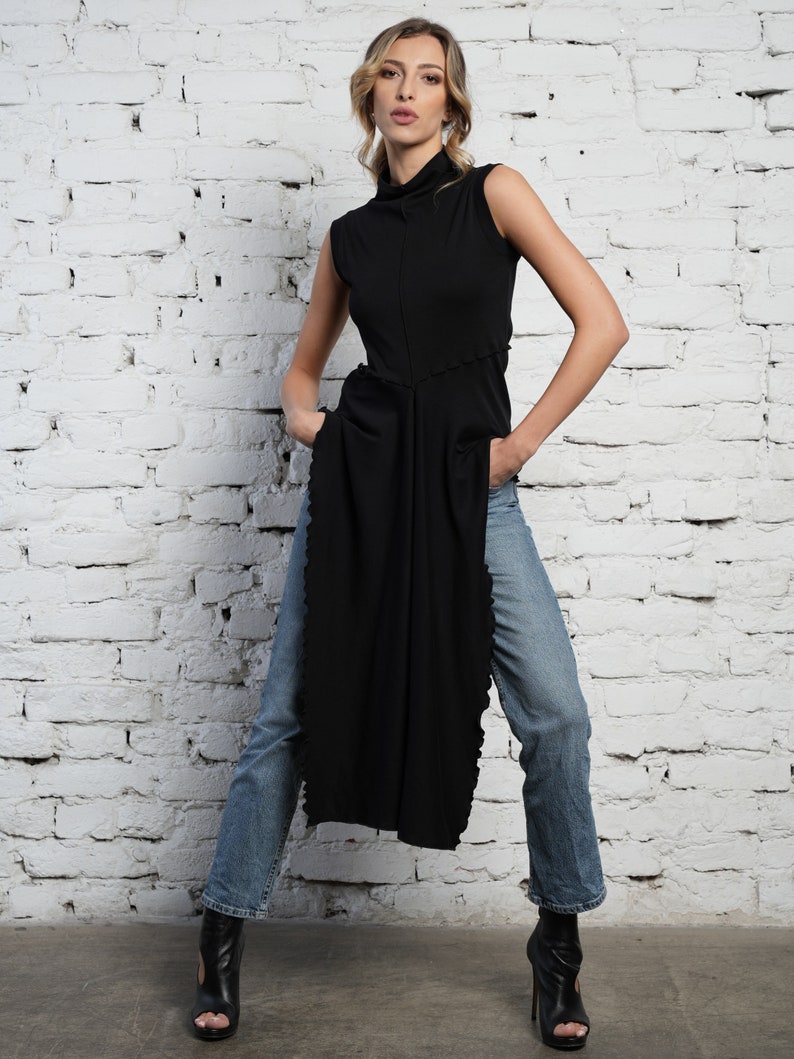 Op de afbeelding: Een zwart shirt met een hoge hals en een lange, asymmetrische zoom. Het shirt wordt gedragen met een blauwe jeans en zwarte hakken.