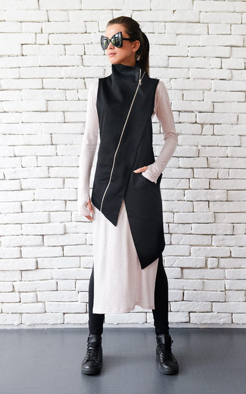 May include: A black sleeveless vest with a diagonal zipper and a long, asymmetrical hemline. The vest is worn over a long-sleeved, light-colored shirt and black pants. The person is wearing black sunglasses and black high-top sneakers.