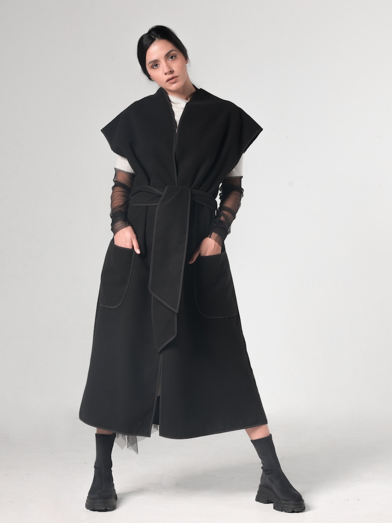 May include: A long black sleeveless coat with a tie belt and pockets. The coat has a contrasting black stitching detail.