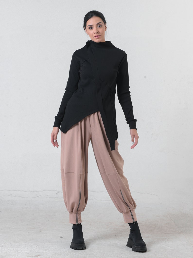 May include: A black mock neck shirt with a long sleeve and a diagonal seam detail. The shirt is paired with loose-fitting, light brown pants with zippered ankles.